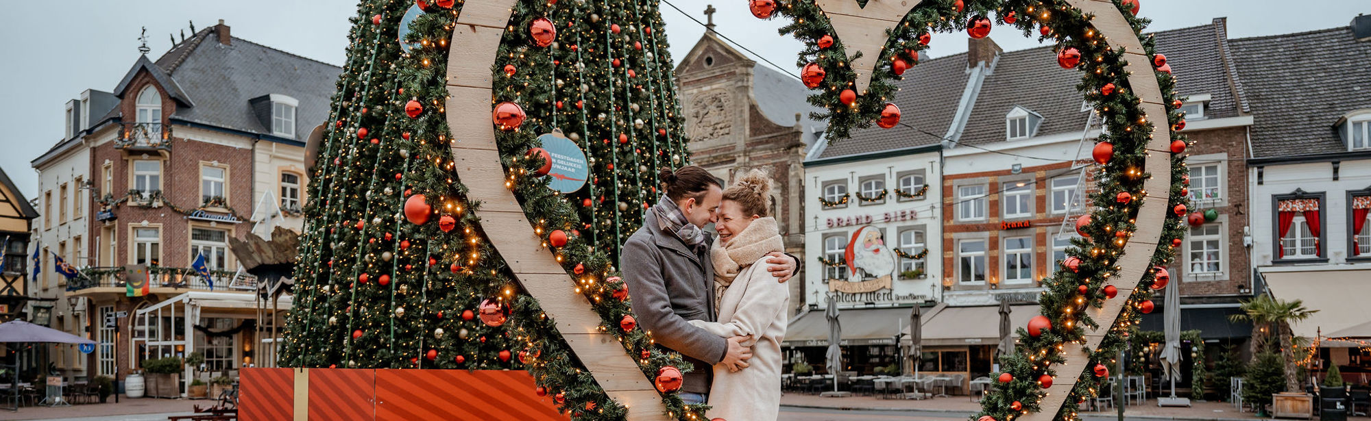 Verliefd koppel knuffelt achter een groot hart op de Markt in Sittard met op de achtergrond een grote kerstboom