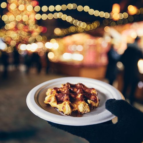 Een hand met handschoen houdt een bordje vast waarop een wafel ligt. Op de achtergrond kerstkraampjes en feestverlichting.